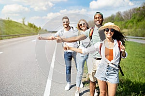 Young multiracial friends standing on highway, showing hitchhiking gesture, stopping car, going on journey together