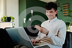 Young multiracial man in shirt smiling while using laptop and credit card on sofa at home