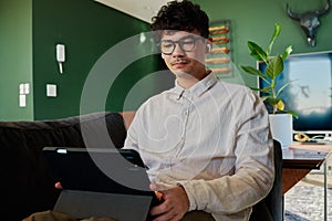 Young multiracial man in shirt using digital tablet with wireless headphones on sofa at home