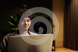 Young multiracial man wearing checkered shirt using laptop on sofa in house at night