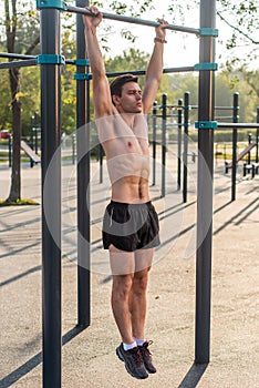 Young muscular athlete doing pull-up exercises hanging with straight arms on a horizontal bar in the park.