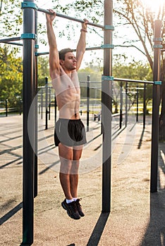 Young muscular athlete doing pull-up exercises hanging with straight arms on a horizontal bar in the park.