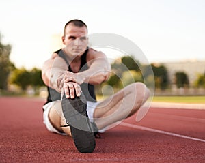 Young muscular athletic runner man stretching and touching his feet on a running court in sitting position before starting of