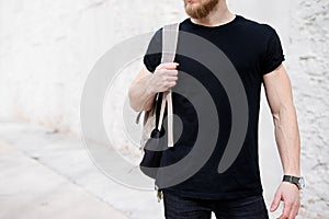 Young muscular bearded man wearing black tshirt and backpack posing outside. Empty white concrete wall on the background