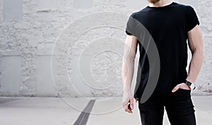 Young muscular bearded man wearing black tshirt and jeans posing outside. White bricks wall on the background