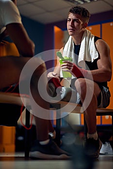 Young muscular boxing player sitting in dressing room holding water bottle facing his coach and talking