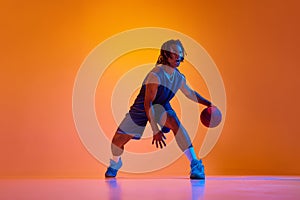 Young muscular man, athlete in motion, playing basketball, doing dribbling against orange background in neon lights