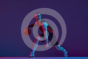 Young muscular man, basketball player in motion, dribbling ball against purple studio background in neon light. Winner