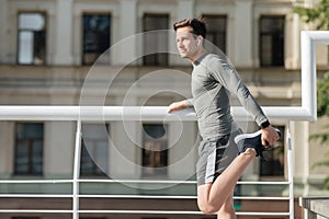 Young muscular man in sportswear and wireless headphones doing stretch for leg