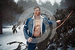 Young muscular man in unbuttoned jacket with bared breast stands next to pine tree in winter forest.