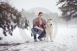 Young muscular man in unbuttoned shirt sits and hugs dog Malamute at walk in winter misty forest.