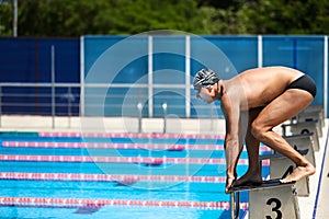 Young muscular swimmer in low position ready to jump on starting block in a swimming pool.