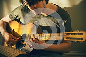 A young musician plays a tune on an old battered guitar, consulting the notes on a sheet of paper that lies on his knee