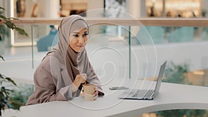 Young muslim businesswoman in hijab sitting in office at desk with laptop stirring coffee in paper cup leisure happy