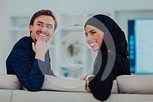 Young muslim couple woman wearing islamic hijab clothes sitting on sofa watching TV together during the month of Ramadan