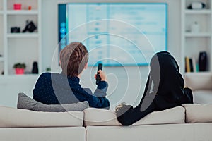 Young muslim couple woman wearing islamic hijab clothes sitting on sofa watching TV together during the month of Ramadan