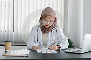 Young muslim female doctor in brown hijab headscarf working with medical papers while sitting in modern clinic office.