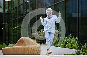 Young Muslim female student outside university campus, happy dancing outside building, woman in hijab listening to music