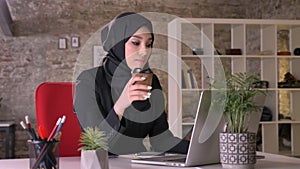 Young muslim girl in hijab is working with laptop in office, drinking coffe, work concept, business concept