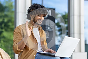 A young Muslim man is talking on a video call on a laptop, sitting outside on a bench in headphones and waving his hand