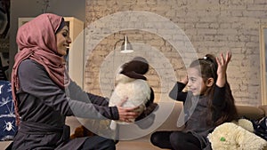 Young Muslim mother in hijab gives her daughter teddy sheep, family sitting on a sofa, idyll, home comfort in the