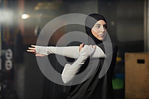 Young Muslim sportswoman in hijab and activewear stretching left arm