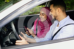 Young Muslim Spouses Riding Car And Using Smartphone Together, Through Window Shot