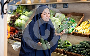 Young muslim woman in abaya and chador choosing cucumbers in store