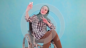 Young muslim woman disabled in a wheelchair takes a selfie with her phone on a blue background