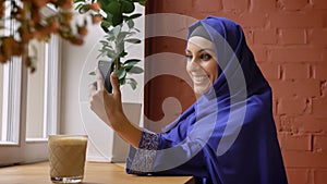 Young muslim woman in hijab having video chat through cell phone, smiling, sitting in modern cafe