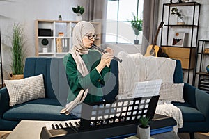 Young muslim woman in hijab plays flute on bright living room at home.