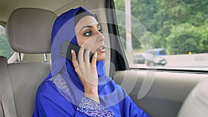 Young muslim woman in hijab sitting in car on passenger rear seat and talking on cell phone
