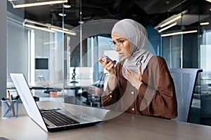 A young Muslim woman in a hijab is sitting at a desk in the office and using an inhaler. He has an asthma attack, holds