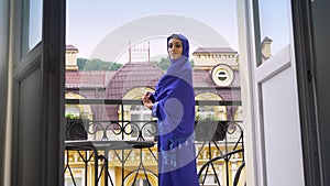 Young muslim woman in hijab standing on balcony, turning and looking at camera, beautiful female