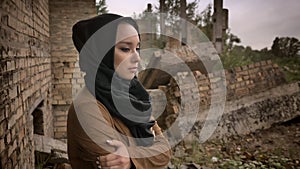 Young muslim woman in hijab standing near ruined building and looking at camera with scared and worried expression, ruin