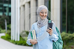 Young muslim woman in hijab walking outside university campus, female student smiling contentedly using app on phone