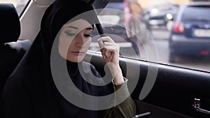 Young muslim woman looking at her phone during the trip by car. Thoughtfully looking on the street through the car
