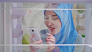 Young muslim woman in a national headscarf looks into an empty refrigerator and orders food at home through an app in