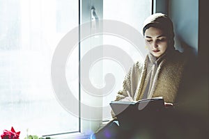 Young Muslim woman sits on the window sill. Middle-eastern writer woman sitting thoughtfully near the window and writing her story