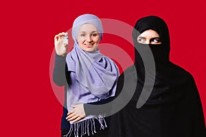 Young Muslim women wearing headscarves. One smiles and holds the vaccine. The second looks to the side on a red