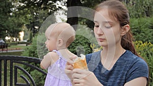 A young nanny picking pieces of a baguette and putting them into her mouth while keeping an eye on a baby girl.