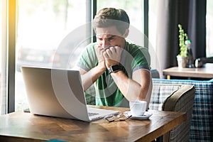 Young nervous businessman in green t-shirt sitting and working on laptop, bitting his nails and looking at screen with worry face