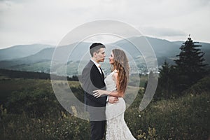 Young newly wed couple, bride and groom kissing, hugging on perfect view of mountains, blue sky