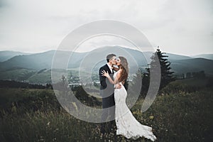 Young newly wed couple, bride and groom kissing, hugging on perfect view of mountains, blue sky