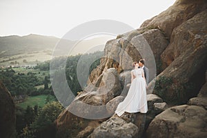 Young newly wed couple, bride and groom kissing, hugging on perfect view of mountains, blue sky