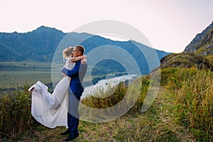 Young newly wed couple, bride and groom kissing, hugging on perfect view of mountains, blue sky