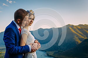 Young newly wed couple, bride and groom kissing, hugging on perfect view of mountains, river and blue sky