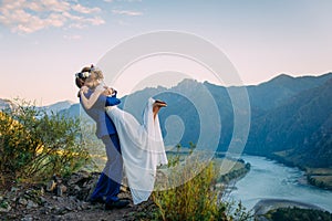 Young newly wed couple, bride and groom kissing, hugging on perfect view of mountains, river and blue sky photo