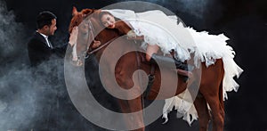 Young newlywed couple, beautiful beauty bride in fashion white bridal wedding costume riding on strong muscular horse standing by