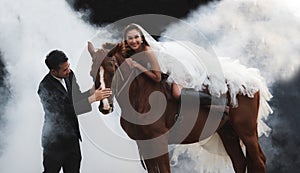 Young newlywed couple, beautiful beauty bride in fashion white bridal wedding costume riding on strong muscular horse standing by
